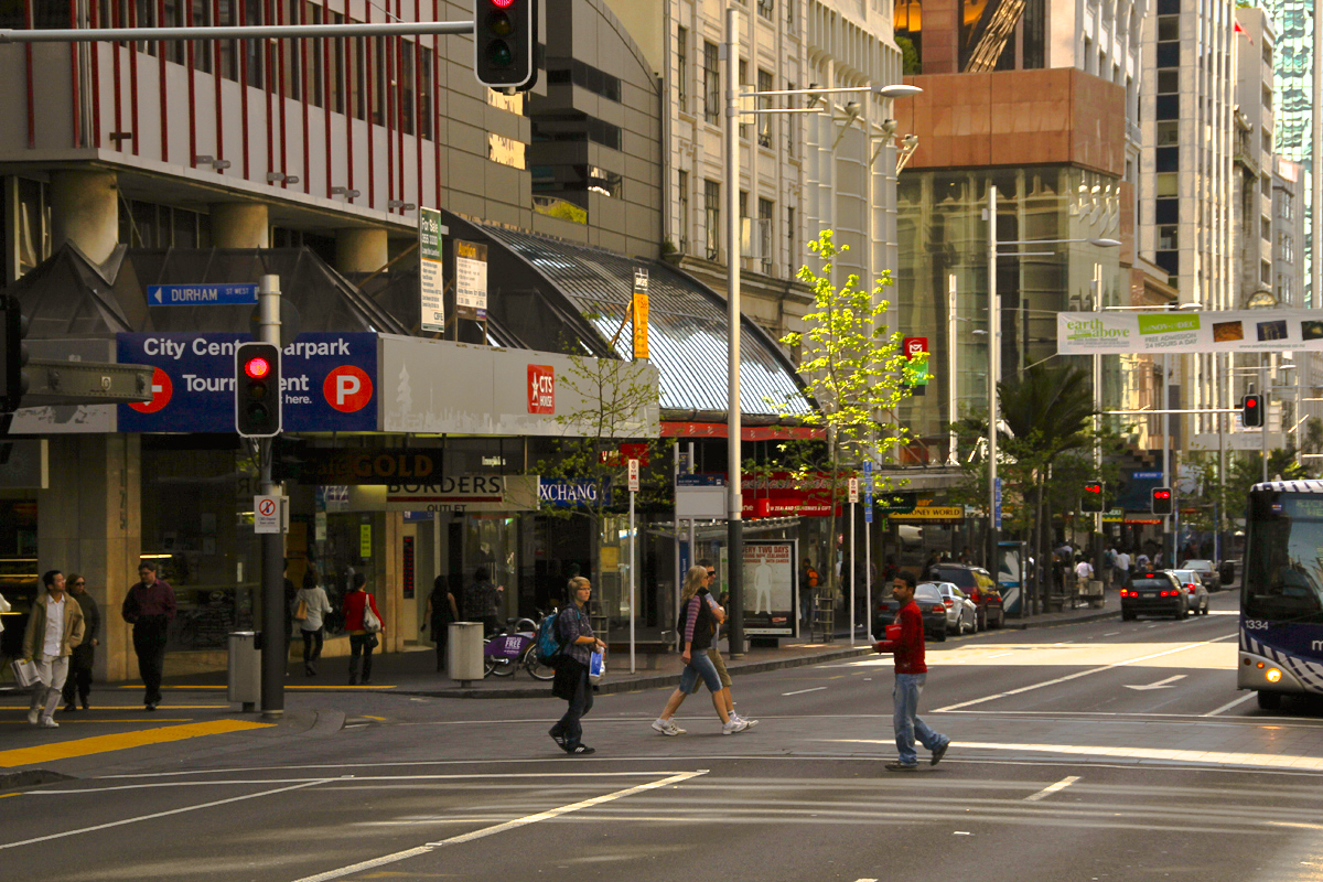 People crossing Queen Street in central Auckland, with shopfronts and a bus along the road.