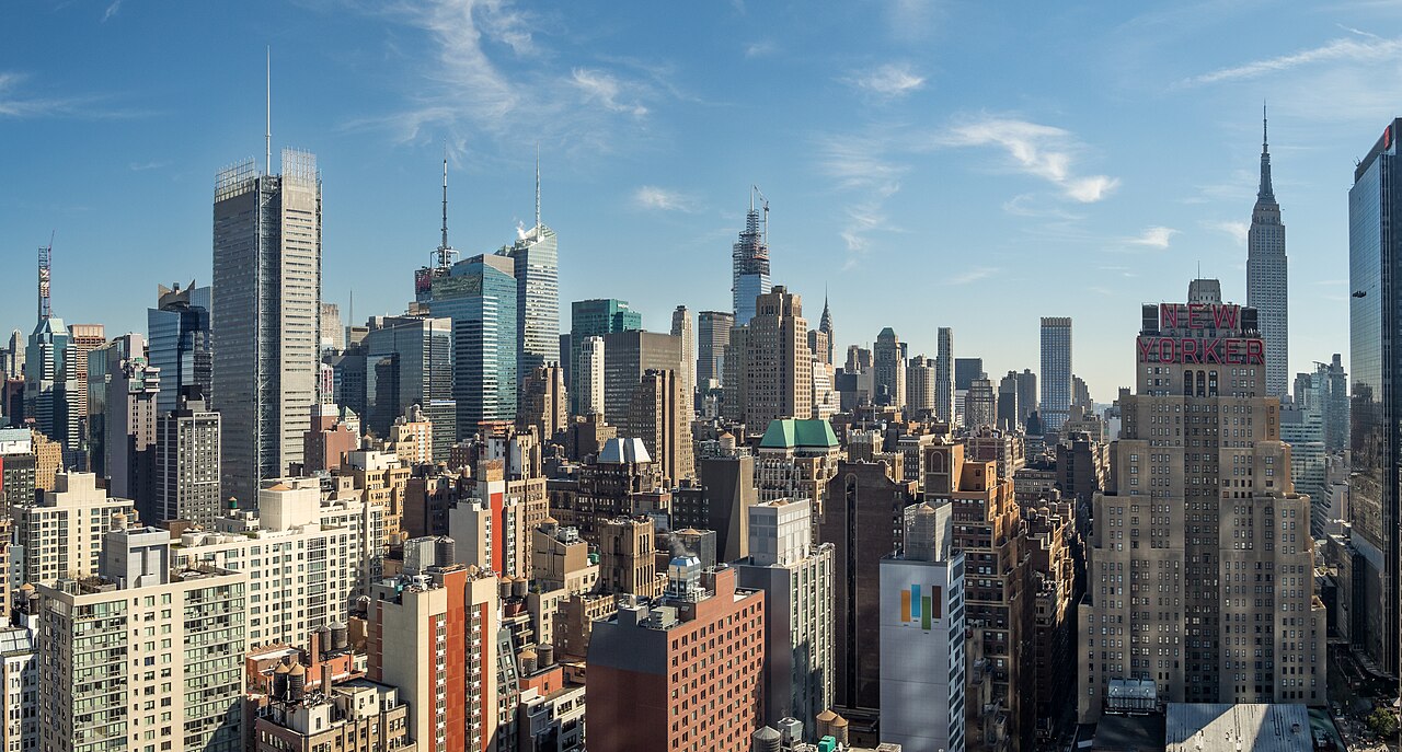 Manhattan rooftops and skyscrapers, including the Empire State Building, under a blue sky.